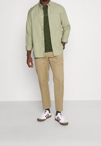 Lys grøn langærmet skjorte over en mørkegrøn t-shirt, parret med beige bukser og hvide sneakers med røde striber og brune såler.