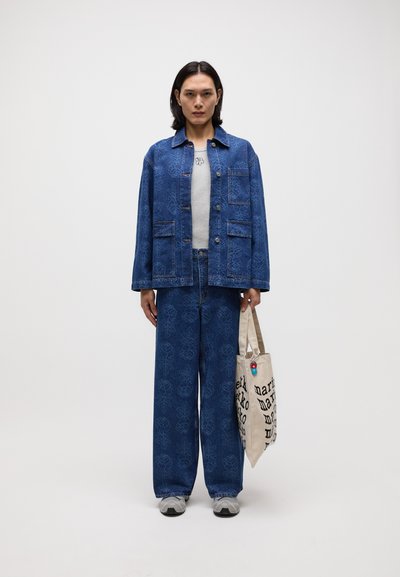 Veste en denim bleu et pantalon large assorti à motif floral. T-shirt gris en dessous. Tient un sac fourre-tout beige avec du texte noir.