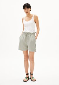 Lichtgrijze satijnen shorts met een elastische tailleband en een trekkoord, gecombineerd met een witte geribbelde tanktop en zwarte sandalen met groene accenten.