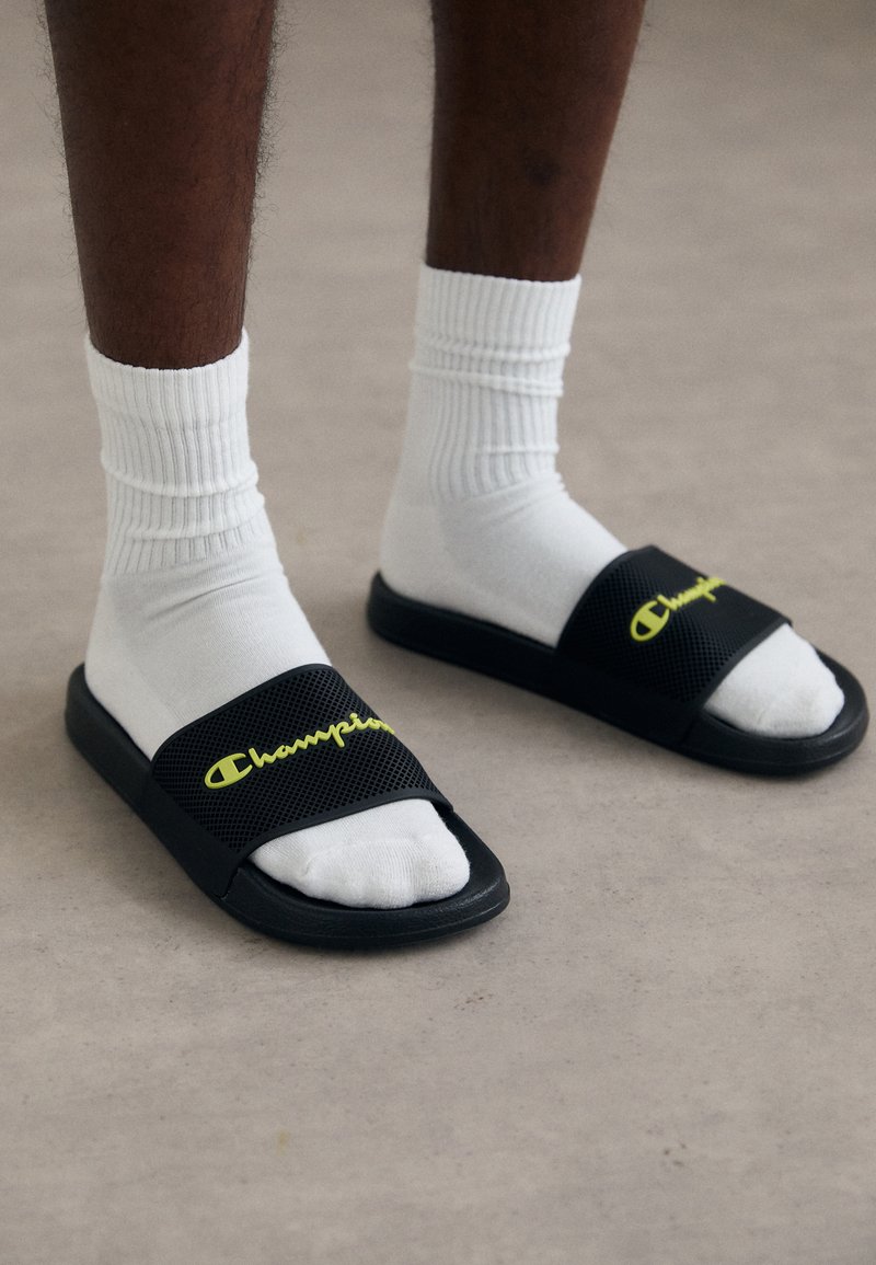 Schwarze Rutschsandalen mit einem strukturierten Riemen, auf dem das gelbe "Champion"-Logo zu sehen ist. Getragen mit weißen gerippten Crew-Socken und auf einer neutralen Oberfläche liegend.