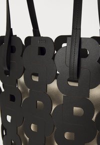 Bolso tote negro con diseño de logo recortado. Presenta una textura mate suave y asas de cuero resistentes. El material interior es una tela tejida ligera.