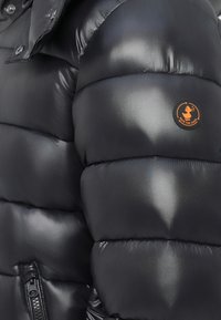 Maniche di giacca nera lucida imbottita con tasca con zip e patch rotondo arancione con la scritta "Save The Duck."