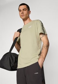 Olivgrünes Baumwoll-T-Shirt mit kurzen Ärmeln, mit einem PUMA-Logo auf der Brust und schwarzen Akzenten entlang der Ärmel. Modell hält eine schwarze Sporttasche.