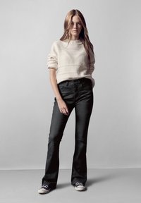 Suéter beige claro con textura horizontal, combinado con jeans negros de campana y zapatillas negras. El modelo se encuentra frente a un fondo neutro.