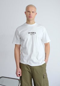 Camiseta blanca de algodón con "DICKIES SINCE 1922" impreso en negro. El modelo lleva pantalones cortos de cargo verde oliva. Diseño minimalista, de mangas cortas.