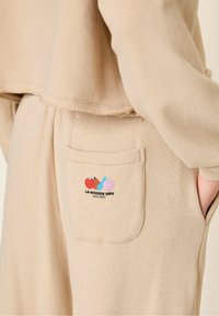 Beige bomulds jogger med en sidelomme; har et flerfarvet broderet logo af to æbler og teksten "LA MAISON AMV siden 2005."