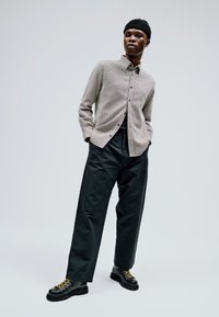 Cămașă texturată gri deschis cu buton, pantaloni negri cu talie înaltă și cizme negre cu șireturi galbene. Modelul poartă o căciulă neagră.