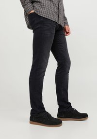 Schwarze Slim-Fit-Jeans aus Denim; verfügt über Seitentaschen, ein elegantes Design und ist kombiniert mit schwarzen Wildlederschuhen mit hellbrauner Sohle.