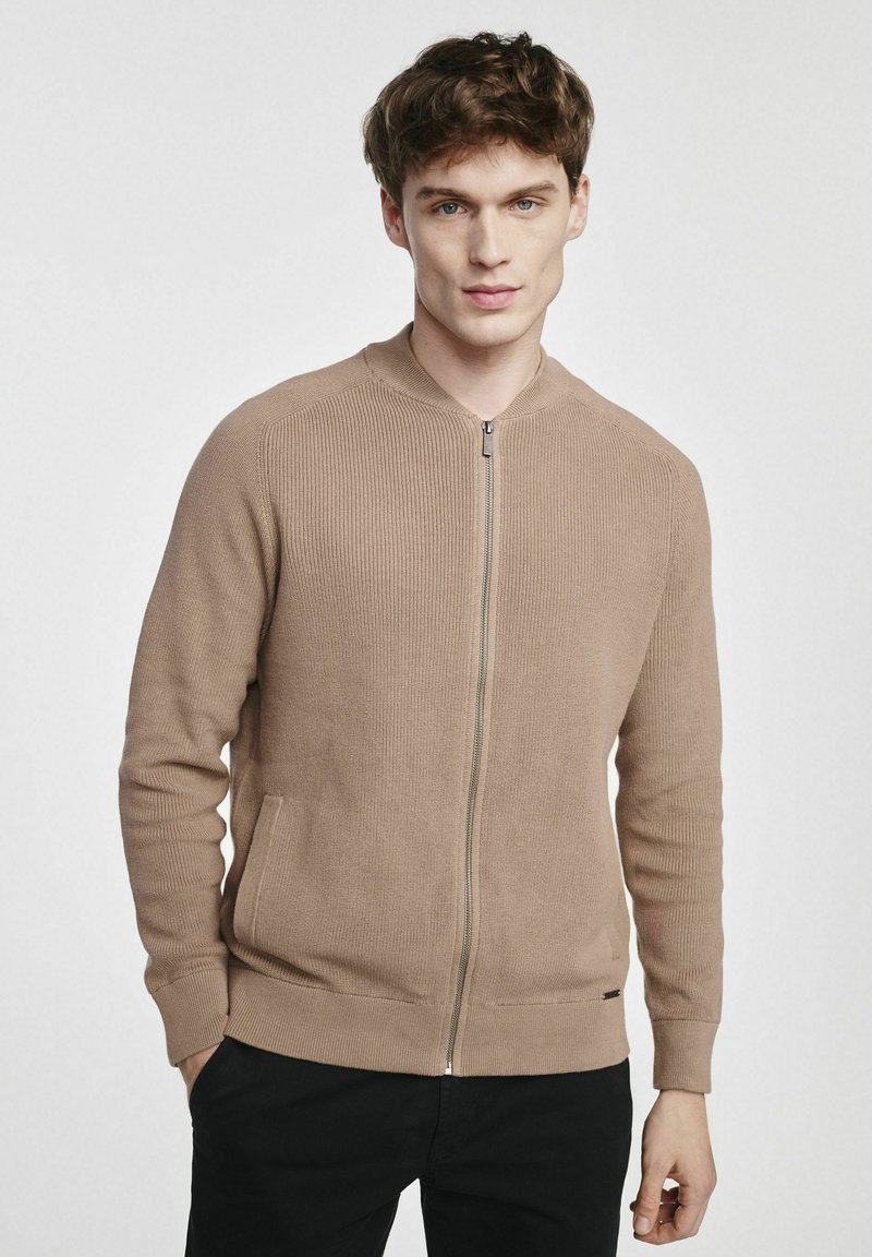 Beżowy dziergany kardigan z zamkiem błyskawicznym, prążkowaną fakturą, bocznymi kieszeniami i okrągłym dekoltem. Dopasowany krój i długie rękawy.