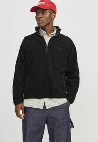 Sort fleece pullover med kvart lynlås og broderet "Originals" logo på brystet. Båret sammen med en ternet skjorte under.
