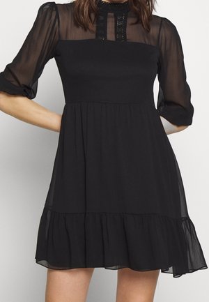 Frau in einem schwarzen, transparenten, langärmligen Kleid mit Spitzenbesatz im Passe und einem gerafften Saum, stehend mit den Händen in die Hüften gestützt.