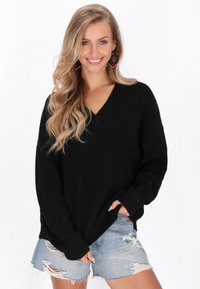 Tânără zâmbitoare, îmbrăcată cu un pulover negru supradimensionat, pantaloni scurți din denim deschis cu zone ruptte și cercei mari roz și negri, având părul lung blond.