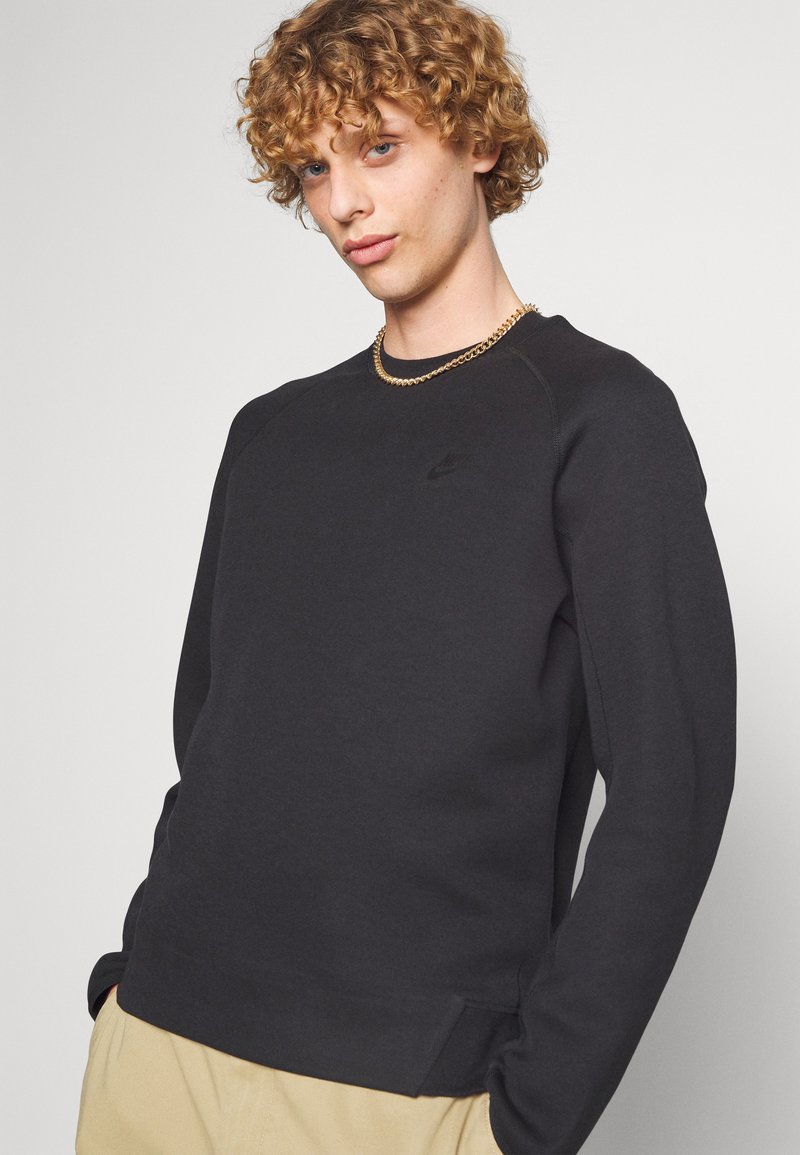 Sweatshirt noir en tissu doux avec un col rond, des manches raglan et un petit logo sur la poitrine. Présente des poignets et un ourlet côtelés.