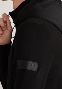 Personne portant un pull noir texturé avec un écusson du logo JOOP! sur la manche, ajustant la fermeture éclair d'une veste noire à capuche à motifs.