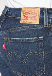 Jeans de mezclilla azul con una etiqueta marrón que presenta un diseño gráfico, costuras amarillas y una etiqueta roja de Levi's en el bolsillo trasero.