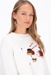 Hvid sweatshirt med en broderet brun gravhund og farverige accenter. Har rød tekst og en ballongrafik. Lange ærmer.