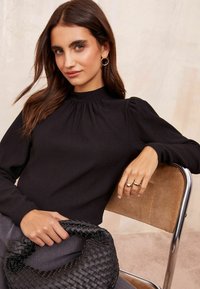 Maglione nero a costine con maniche a sbuffo, collo rotondo e tessuto texture. Il modello tiene in mano una borsa nera intrecciata ed è seduto su una sedia di metallo.