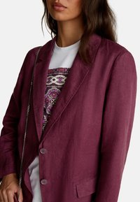 Femme portant un blazer violet foncé sur une chemise blanche à motifs, tenant une petite pochette rouge avec une bandoulière en chaîne argentée.