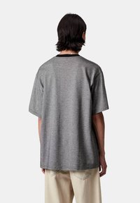 Osoba z włosami średniej długości w ciemnym kolorze nosi luźną szaro-skalną koszulę z krótkim rękawem oraz jasne beżowe spodnie, stojąc tyłem na jednolitym tle.