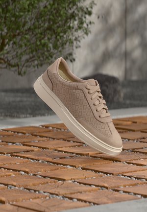 Baskets beige avec des côtés à motifs et une semelle blanche flottant au-dessus d'un sol en carrelage terracotta avec des cailloux, un buisson vert et un mur gris en arrière-plan.