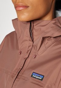 Chaqueta impermeable marrón con cuello alto, que presenta toggles ajustables y un logo de Patagonia en el pecho. Textura suave y mate.