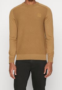 Stickad tröja i kamelfärg med ribbad struktur, långa ärmar, rundad hals och ett litet fyrkantigt logopatch på bröstet.