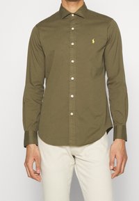 Camicia a maniche lunghe verde oliva con colletto a scorrere, bottoni bianchi e un piccolo logo giallo sul lato sinistro del petto. Tessuto dalla consistenza liscia.