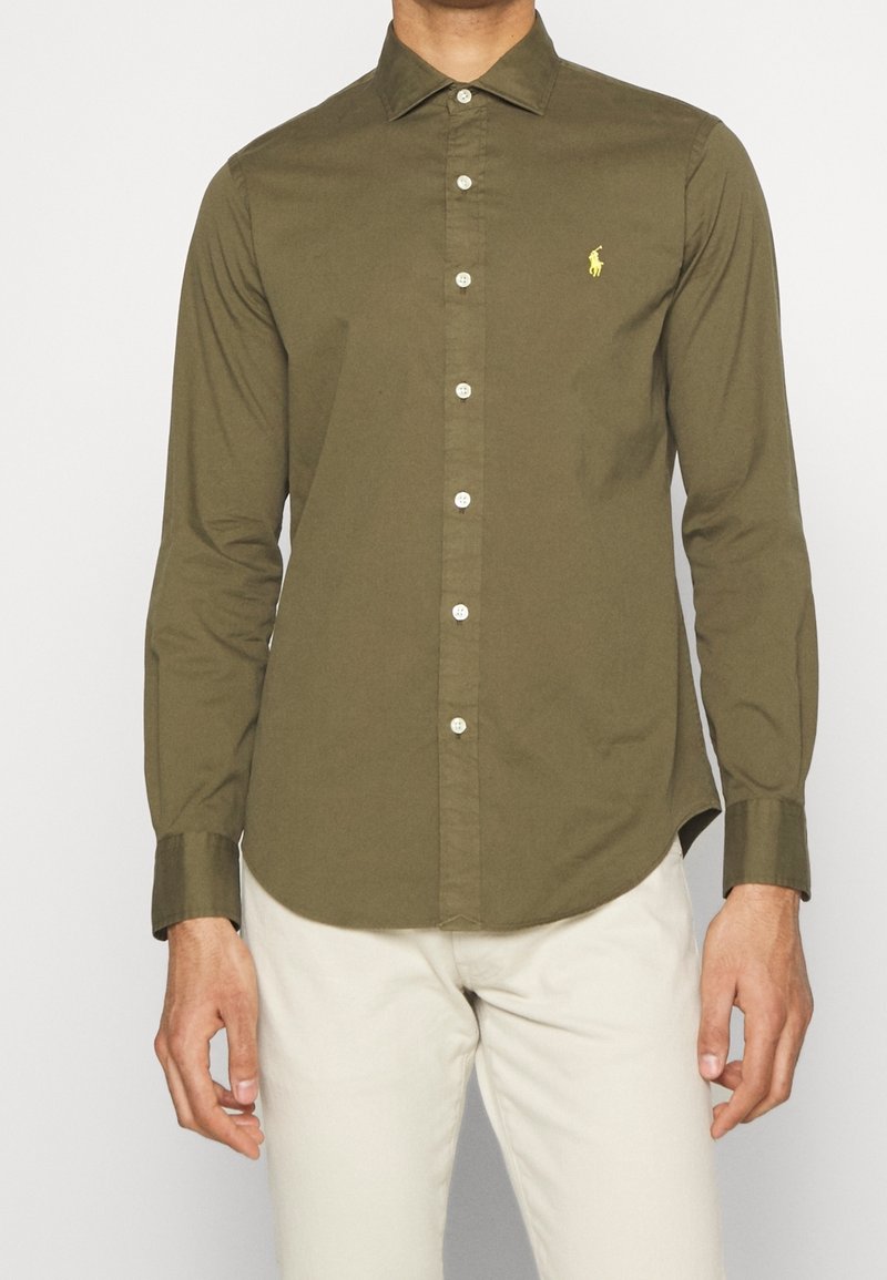 Camicia a maniche lunghe verde oliva con colletto a scorrere, bottoni bianchi e un piccolo logo giallo sul lato sinistro del petto. Tessuto dalla consistenza liscia.