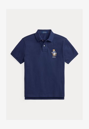 Polo majica mornarsko plave boje s dva gumba i vezanim medvjedićem u blejzeru koji sjedi na stolcu iznad riječi „POLO“ na prsima.