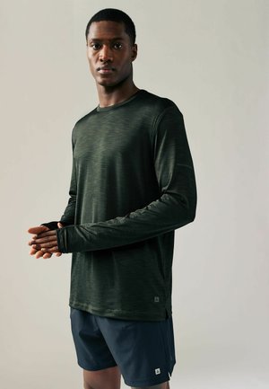 Donkergroen sportshirt met lange mouwen en een gestructureerd oppervlak, voorzien van duimopen en een relaxte pasvorm, gecombineerd met marineblauwe shorts.