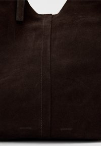 Saco de tote em camurça castanha com textura suave, topo em formato U e costura branca subtil ao longo da costura. Logotipo em relevo na parte inferior.