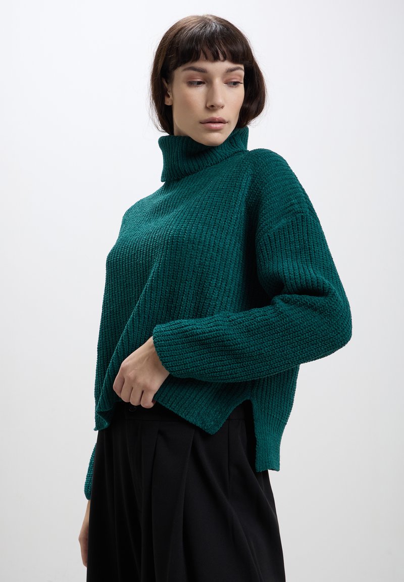 Pull en tricot vert avec un col montant, en tissu texturé et design à épaules tombantes. Porté sur un pantalon large noir.