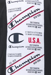 Musta kangas, jossa on suuri valkoinen etiketti, jossa on mustaa ja punaista tekstiä. Siinä on brändin nimi "Champion" ja "U.S.A." koristeilla.