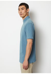 Light blue short-sleeve polo shirt made of textured cotton with a buttoned collar, featuring side slits at the hem for ease of movement.
