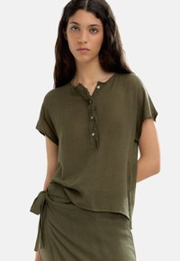 Oliwkowy top z krótkimi, podwiniętymi rękawami, z postrzępionym dekoltem i trzema guzikami. Wykonany z lekkiego materiału.
