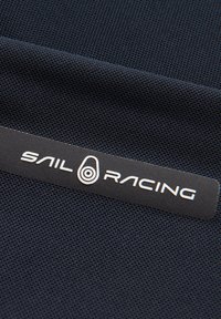 Marineblauter Stoff mit einer netzartigen Textur, versehen mit einem schwarzen Etikett, auf dem in weißen Buchstaben "SAIL RACING" steht, sowie einem ovalen Logodetail.