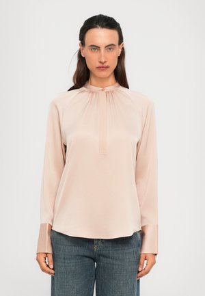 Kvinne med langt, mørkt hår iført en lys rosa langermet bluse med høy krage og blå jeans, står mot en lys, ensfarget bakgrunn.