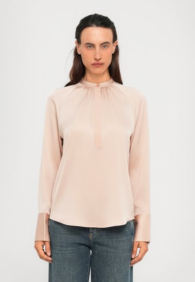 Vrouw met lang donker haar, gekleed in een lichtroze blouse met lange mouwen en hoge kraag, en blauwe jeans, staand tegen een effen lichte achtergrond.