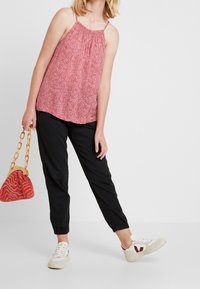 Débardeur à motif floral rouge avec bretelles ajustables, associé à un pantalon noir fuselé et des baskets beiges. Tient un sac à main texturé rouge.