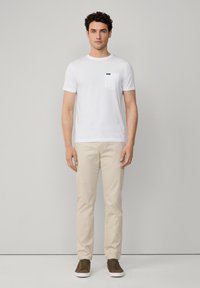 T-shirt bianca in cotone con taschino, chinos beige e sneakers marroni. Il modello è in piedi contro uno sfondo neutro.