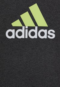 Szara bawełniana koszulka z białym logo "adidas" i trzema geometrycznymi paskami w jaskrawym limonkowym kolorze. Miękka tekstura, standardowy krój z okrągłym dekoltem.