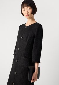 Abito in tweed nero con scollo rotondo, maniche a tre quarti e bottoni decorativi. Presenta tasche e una finitura testurizzata. Tiene in mano una pochette nera.