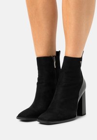 Bottines noires en daim avec fermeture éclair, présentant un bout pointu et un talon épais, mettant en valeur un accent en cuir lisse.