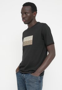 Μαύρο βαμβακερό T-shirt με στρογγυλή λαιμόκοψη και ορθογώνιο μπεζ σχέδιο με το κείμενο "JACK & JONES" σε λευκό. Συνδυάζεται με μπλε τζιν παντελόνι.