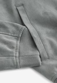 Gros plan d'une manche de sweat-shirt grise avec poignet côtelé et détail de poche verticale sur tissu doux.