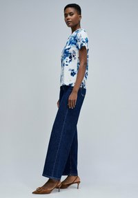 Top floral en blanco con patrones azules, combinado con jeans de pierna ancha en azul marino y tacones marrones. Mangas cortas, corte relajado y costuras visibles.
