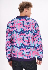 Giacca bomber colorata con un motivo mimetico nei toni del rosa, blu e viola. Il materiale è liscio, con polsini e colletto a coste, e una vestibilità comoda.