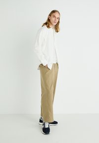 Chemise blanche à boutons, pantalon beige à plis et baskets marines avec accents blancs. Le modèle se tient devant un fond uni.