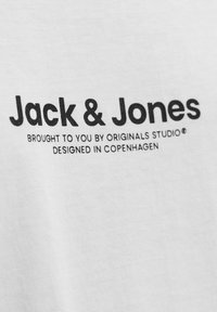 Balta medvilninė marškinėlių su juodu tekstu, kuris sako „Jack & Jones“, o po juo pateikiama papildoma mažesnė informacija apie originalią studiją ir dizaino detales.