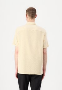 Beige Kurzarmhemd aus strukturiertem Stoff mit Kragen, ausgestattet mit einem einzelnen Rückenschlitz und einem geraden Saum. Kombiniert mit schwarzen Hosen.